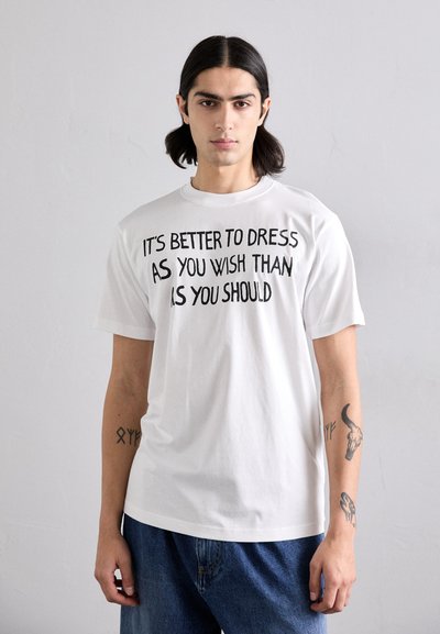 T-shirt bianco in cotone con scollo tondo, caratterizzato da un testo nero audace che recita: "È MEGLIO VESTIRSI COME DESIDERI CHE COME DOVRESTI."