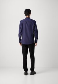Cămașă albastră cu nasturi, dintr-un material combinat cu bumbac, cu un model geometric, mâneci lungi și guler rotunjit, purtată cu pantaloni negri și pantofi.