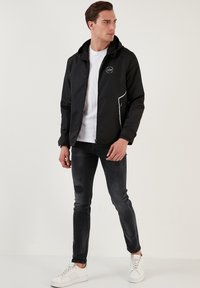 Jachetă neagră cu glugă, cu margini albe, buzunare cu fermoar; combinată cu un tricou gri deschis cu nervuri, blugi negri deteriorați și adidași albi.