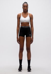 Γυμνασμένη γυναίκα στέκεται μπροστά με άσπρο αθλητικό σουτιέν, μαύρο σορτς Under Armour, μαύρες κάλτσες και μαύρα αθλητικά παπούτσια σε απλό φόντο.