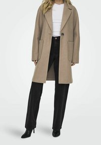 Abrigo de lana beige con solapas de muesca, un solo botón y dos bolsillos frontales; combinado con jeans negros de cintura alta y tacones puntiagudos.