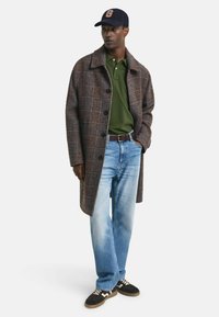 Man in een bruine geruite jas, groene polo, lichtblauwe spijkerbroek, zwarte sneakers en een marineblauwe pet met een "G" embleem, staand met één hand in zijn zak.