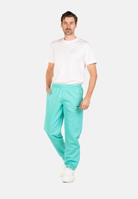 Een model draagt een wit T-shirt en turquoise joggers met een elastische tailleband, die een zachte textuur en taps toelopende boorden hebben.