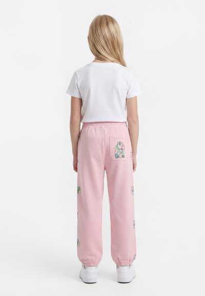 Hosszú szőke hajú gyermek áll háttal, fehér pólót, rózsaszín virágmintás nadrágot, rugalmas szegélyű bokarészt és fehér tornacipőt visel.