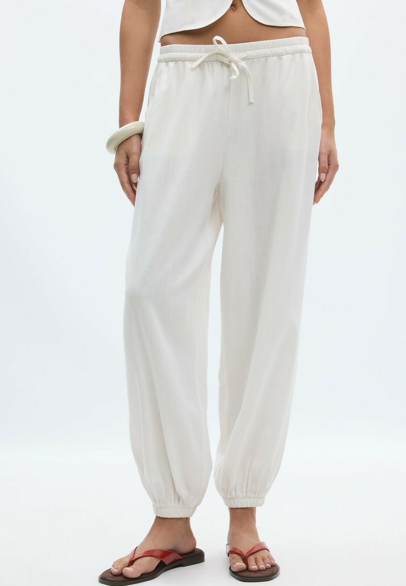 Femme portant un pantalon de jogging blanc avec cordon de serrage et poignets élastiques, un top court blanc, un bracelet au poignet gauche et des sandales rouges.