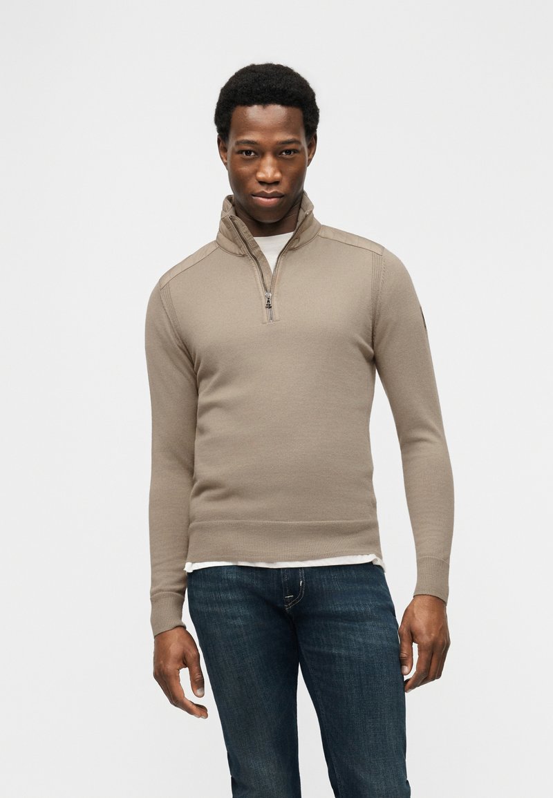 Άνδρας που φοράει μπεζ πουλόβερ με φερμουάρ στο μισό μήκος πάνω από λευκό πουκάμισο και σκούρο μπλε τζιν, στεκόμενος μπροστά σε απλό ανοιχτό γκρι φόντο.