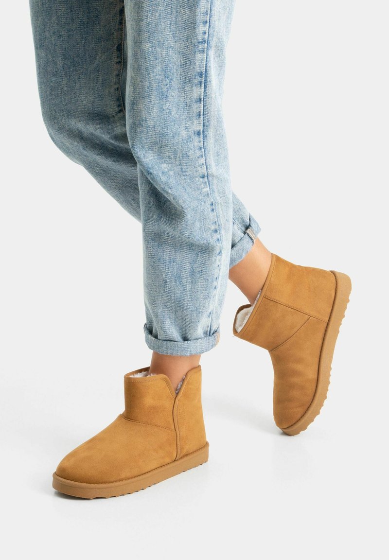 Personne portant un jean bleu clair retroussé et des bottines en suède camel avec doublure en polaire, debout sur une surface blanche.