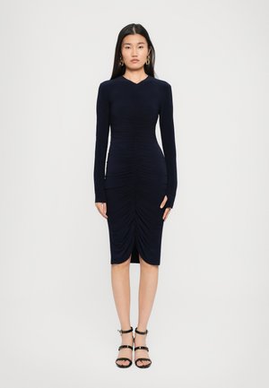 Woman wearing a fitted navy ruched long-sleeve midi dress and black strappy heels, standing against a plain white background.