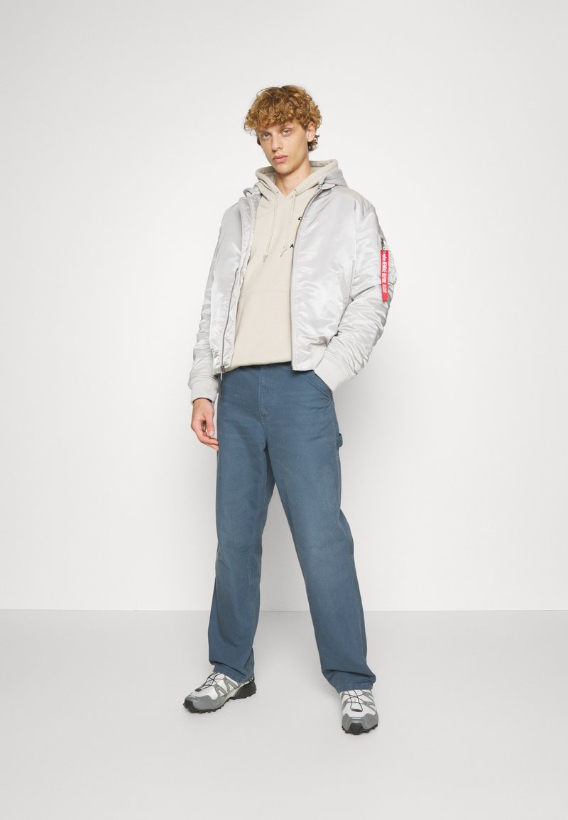 Veste bomber argentée sur un sweat à capuche beige, avec un pantalon bleu et des chaussures grises. La veste présente une accent rouge et des poignets côtelés.