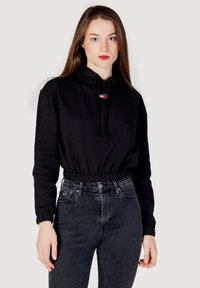 Felpa nera con cappuccio, dotata di orlo e polsini elasticizzati, caratterizzata da un patch logo in rosso, bianco e blu. Indossata con jeans scuri a vita alta.