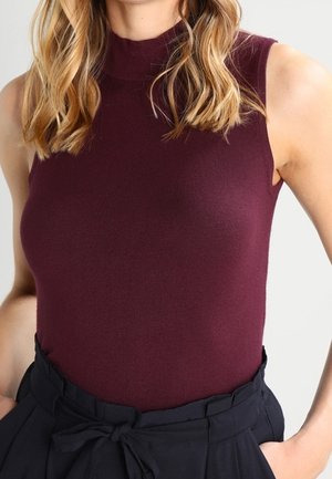 Vrouw met een mouwloze, hooggesloten bordeauxrode top, ingestopt in zwarte, hooggetailleerde broek met een gestrikte riem.