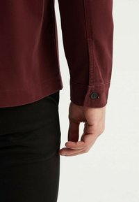 Chemise bordeaux à manches longues en tissu texturé, avec un poignet boutonné et un ourlet droit, associée à un pantalon noir.