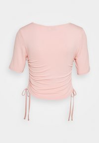 Růžový top s krátkým rukávem a kulatým výstřihem, s detailem na bočních stranách. Materiál vypadá měkký a elastický, s nastavitelnými tkanicemi na spodním lemu.
