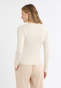 Beżowa bluzka z długim rękawem o dopasowanym kroju i prążkowanej fakturze, zestawiona z jasnymi spodniami w paski z bocznymi kieszeniami.