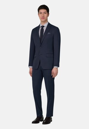 Uomo che indossa un abito blu scuro, camicia a righe, cravatta blu navy, fazzoletto da taschino a motivo e scarpe eleganti nere, in piedi su uno sfondo bianco uniforme.