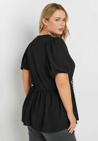 Top nero con maniche a palloncino, vita arricciata e design peplum fluido, caratterizzato da una morbida texture del tessuto. Nessun motivo o accessorio visibile.