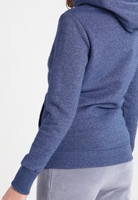 Sininen fleece-huppari, jossa on teksturoitu kangas, huppu, resorit hihansuissa ja helmassa. Selässä on hillitty design-elementti lähellä kaulusta.