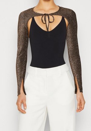Femme portant un pantalon blanc taille haute et un haut noir sans manches avec de longues manches boléro marron scintillantes nouées au niveau du col.