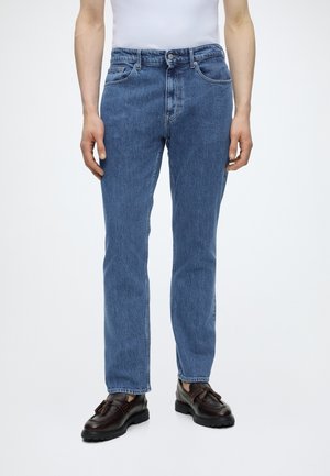 Mand iført blå jeans med lige ben, hvid skjorte og mørkebrune læder tassel loafers, stående mod en ensartet hvid baggrund.