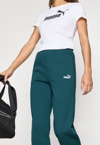 White t-shirt with black "PUMA" logo, paired with teal joggers featuring a small white logo. Model holds a black bag. Simple and sporty design.