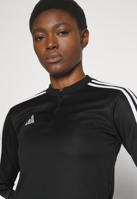 Čierny atletický top s prieh ľadným golierom, priliehavým strihom a bielymi trojpruhovými akcentami na rukávoch. Mäkka, ľahká tkanina.