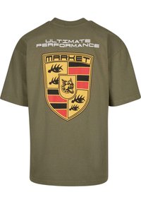 Oliven grøn t-shirt med et stort skjoldgrafik på ryggen, der har sorte og gule accenter. Teksten siger "ULTIMATIV YDEEVNE."