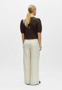 Femme avec une queue de cheval blonde portant un pull marron à manches courtes avec une broderie florale blanche dans le dos et un pantalon large crème.