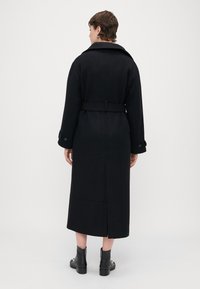 Garšs melns mētelis ar plašu apkakli, jostotu vidukli un sānu šķēlumu; izgatavots no teksturēta auduma. Valkāts ar melnām potītes zābakiem.