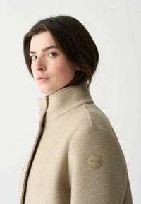 Beige ullblandad jacka med hög krage, med en subtil textur och en cirkulär logotyp-patch på vänster axel.