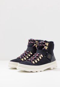 Un paio di scarpe da trekking in suede nero con suole bianche, lacci a motivo rosso e nero, occhielli dorati e cinghie alla caviglia nere su una superficie bianca.