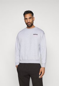 Felpa grigia chiaro con colletto a girocollo, polsini e fondo a costine. Presenta il logo "JUVENTUS" in nero e rosa sulla parte sinistra del petto.