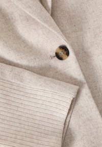 Manteau en laine beige avec un bouton en écailles de tortue et un poignet en tricot côtelé sur la manche.