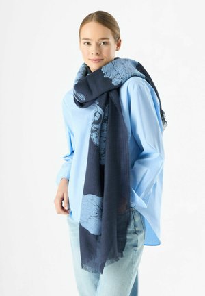 Écharpe bleue à motifs en matériau léger, présentant un design abstrait en bleu plus foncé, drapée autour du cou. Bords à franges visibles.