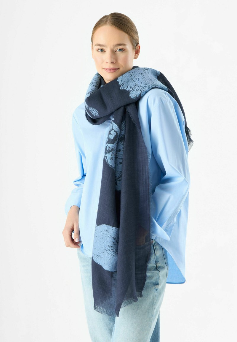 Écharpe bleue à motifs en matériau léger, présentant un design abstrait en bleu plus foncé, drapée autour du cou. Bords à franges visibles.