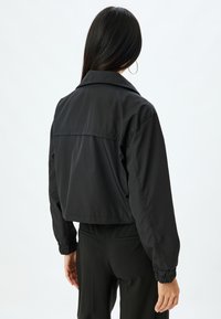 Kraftig svart jacka med krage, elastiska ärmslut och en enda bakstycke-detalj. Slät, lättviktig tyg.