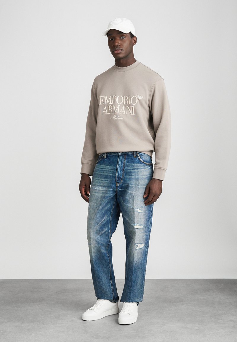 Sweatshirt beige clair avec logo "Emporio Armani", associé à un jean délavé bleu avec déchirures et des baskets blanches. Modèle portant une casquette blanche.