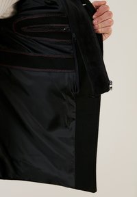 Interior de chaqueta negra con un forro textil suave, múltiples bolsillos con cremallera y acentos de costura roja a lo largo de las costuras.