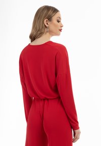 Rød, langærmet jumpsuit med en afslappet pasform og rund halsudskæring. Stoffet ser glat og let ud med et bælte ved taljen for at give form.