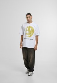 Hvid bomulds t-shirt med et stort gult smiley-ansigt grafik og teksten "Produkt af Internettet" i sort, parret med mørke bukser.