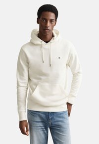 Cream hoodie made of soft fabric, featuring a front pocket, drawstring hood, and small logo. Worn with blue jeans.