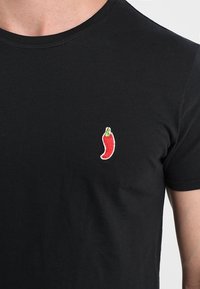 Black cotton t-shirt with short sleeves, featuring a small red chili pepper embroidery on the left side of the chest.