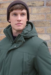 Grüne gesteppte Jacke mit Reißverschluss und Knopfverschluss, ausgestattet mit hohem Kragen und verstellbarer Kapuze, kombiniert mit einer braunen Strickmütze.