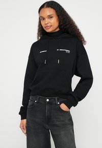 Sweatshirt preta com capuz com bolso canguru, cordão no capuz e logótipo em texto branco. Combinada com jeans pretos de cintura alta, apresentando uma textura subtil.