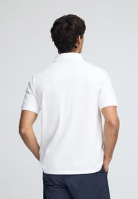 Witte polo met korte mouwen, klassieke kraag, gladde textuur en een losse pasvorm, gezien vanaf de achterkant. Geen zichtbare logo's of patronen.