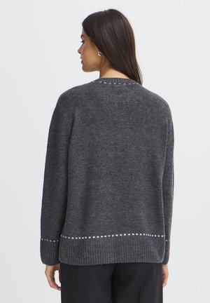 Vrouw met lang donker haar die een donkergrijze gebreide trui draagt met witte stikseldetails bij de zoom en manchetten, van achteren gezien.