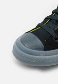 Czarne płócienne buty z gumowym noskiem, akcentowane neonowo żółtym przeszyciem. Posiadają chropowatą niebieską gumową podeszwę i czarne sznurowadła.