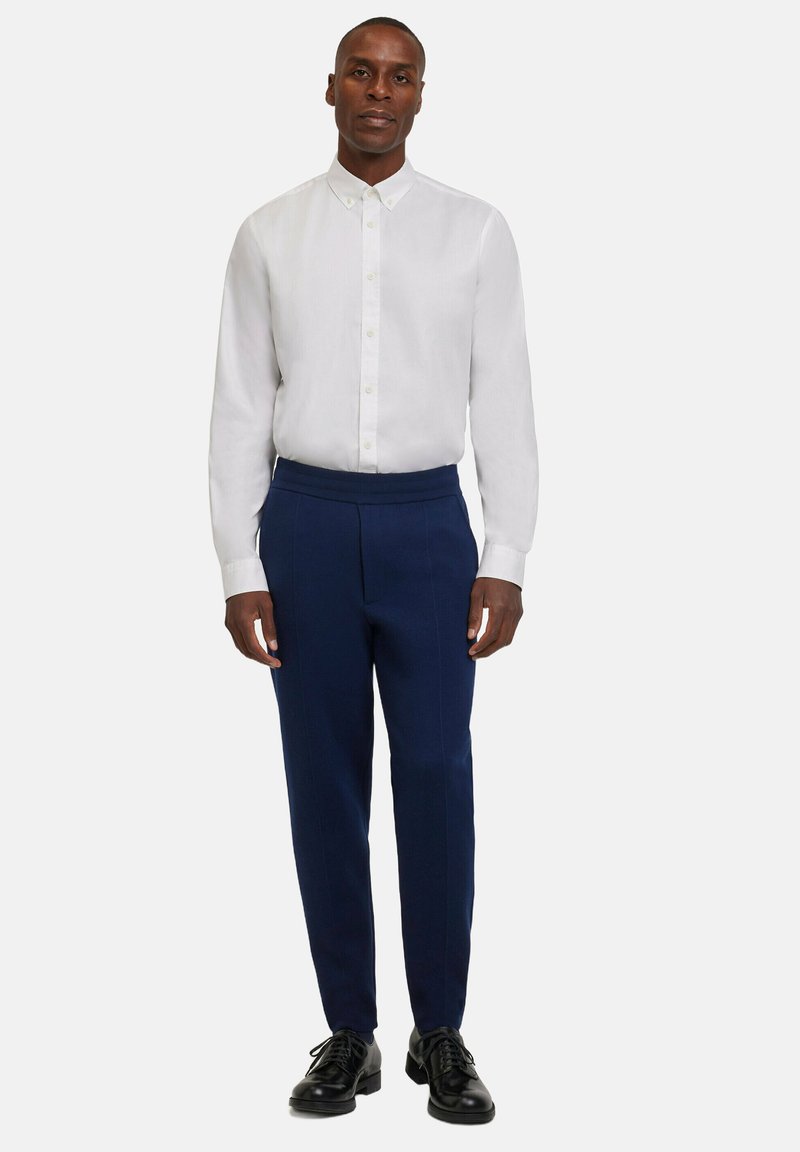 Mann steht mit einem weißen Hemd mit Knöpfen, einer marineblauen Hose und schwarzen Schnürschuhen vor einem schlichten weißen Hintergrund.