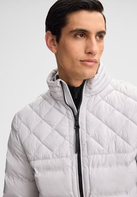 Hellgraue, gesteppte Pufferjacke mit hohem Kragen, Frontreißverschluss und schwarzen Akzenten. Glatte Textur und wattiertes Design für Wärme.
