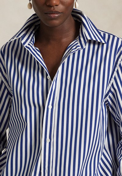 Camicia a righe bianche e blu con bottoni, ampio colletto e tessuto liscio. Presenta strisce verticali e un design dal taglio morbido.
