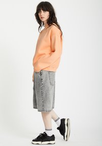 Sweatshirt oversized pêche avec des épaules tombantes, associé à un short en denim gris Bermuda et des baskets noires. Chaussettes rayées blanches visibles.